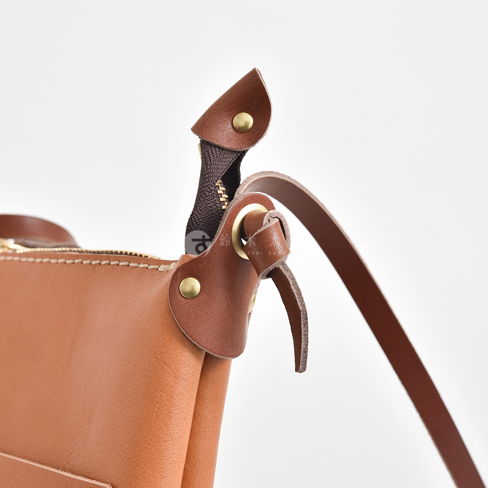 棕色皮革直式隨身單肩包，特寫拉鍊及皮革提把連接處，帶有金色五金配件。