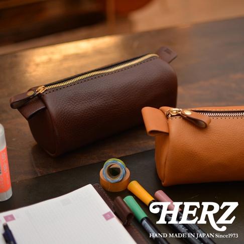 職人手作皮革多用途收納筆袋，一個為深棕色，一個為淺棕色，均採用帶有紋理的皮革材質，配有金色拉鍊和皮革拉環，放置於桌面，旁邊有筆記本、馬克筆和膠帶。