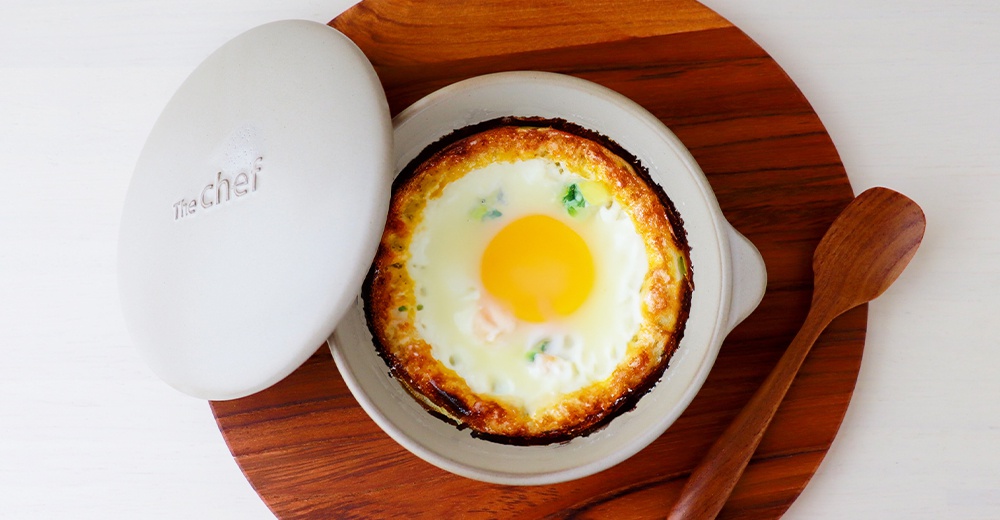 The Chef 品牌白色耐熱陶製圓鍋，內含烘烤完成的雞蛋，蛋黃金黃，蛋白凝固，鍋緣帶有烤色。一旁有木製湯匙，置於木質餐盤上。