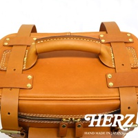 職人手作皮革大型三用旅行後背包，展示其圓潤的頂部提把、兩側的皮革固定帶，以及底部雙拉鍊設計。整體為溫暖的棕褐色，材質為皮革，細節處可見精緻的縫線與金屬卯釘。