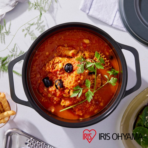 IRIS OHYAMA 萬用無水調理鍋內盛裝著以番茄為基底燉煮的牛肚料理，上方點綴著橄欖和香料，鍋旁擺著烤麵包棒。