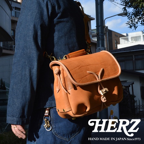 咖啡色皮革製成的職人手作鹿角扣繩郵差包，包身有自然壓紋，搭配淺棕色皮革滾邊與提把，金色金屬釦環與鹿角造型拉繩，側面有鉚釘裝飾。