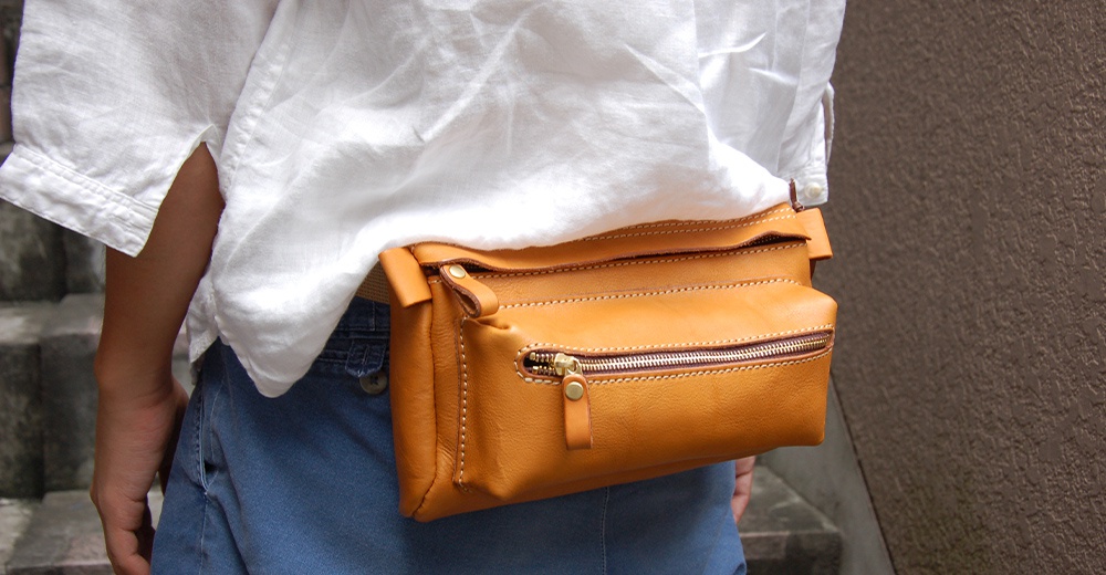職人手作皮製兩用腰掛斜背包，橘棕色皮革，帶有黃銅色拉鍊和縫線細節，可掛於腰間或斜背。
