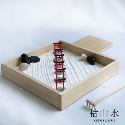 日式枯山水模型，木製方形盆器內鋪有白色細沙，沙上排列著七座紅色鳥居，盆器兩側擺放著黑色及白色鵝卵石，右側有一支木柄小耙子。