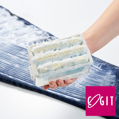 手持一個透明塑膠盒，內含藍色海綿和白色刷毛，盒上方充滿白色泡沫。產品置於藍色牛仔褲上方，牛仔褲上有水漬和泡沫。