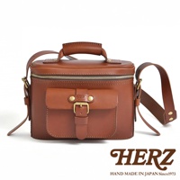 HERZ 經典款棕色皮革相機包，配有頂部提把、可調式肩帶和前置翻蓋口袋，口袋上飾有復古皮帶扣和 HERZ 品牌標誌，下方印有「HAND MADE IN JAPAN Since1973」字樣