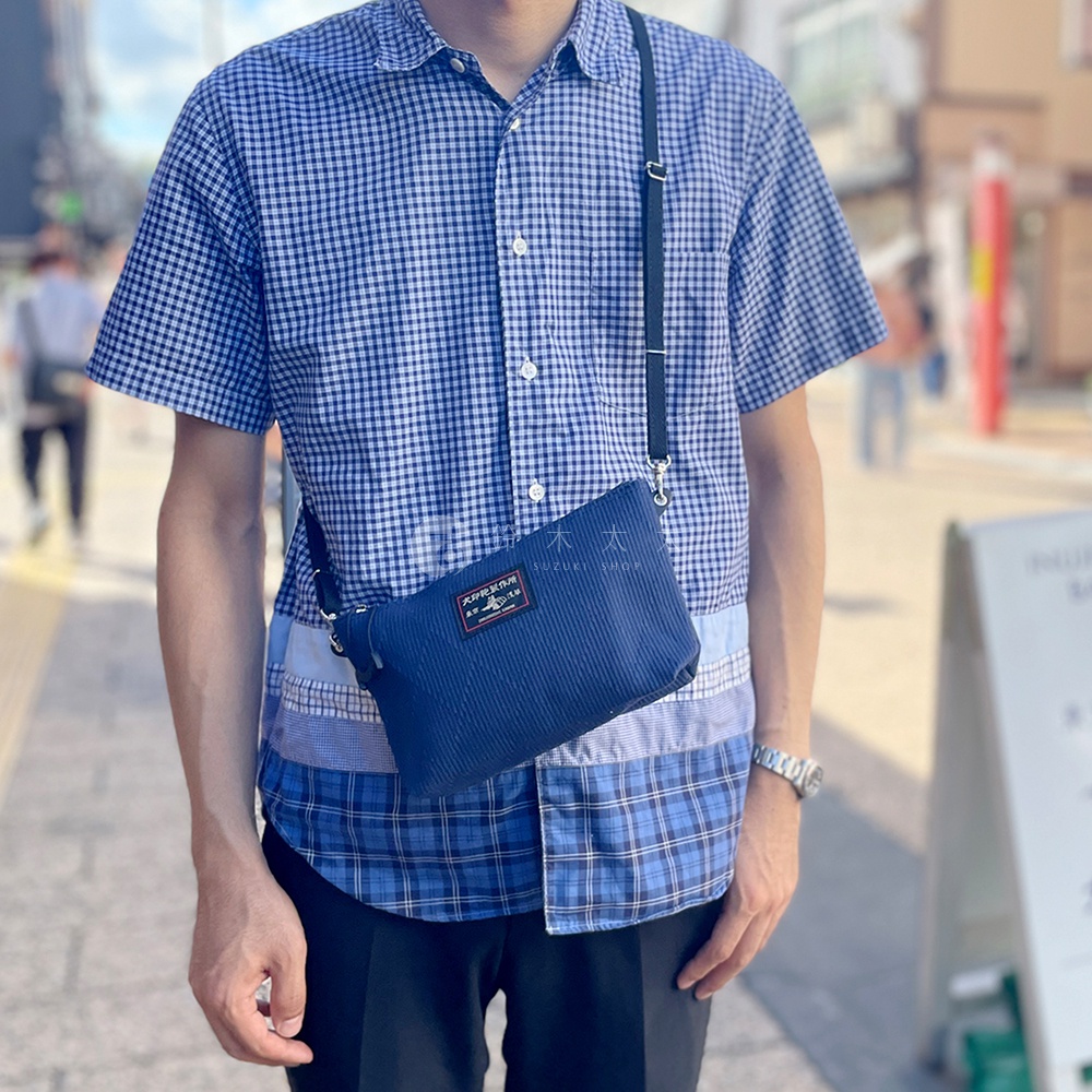 深藍色燈芯絨材質斜背包，正面有紅色標籤，黑色細肩帶，展示於身穿格紋襯衫的男士身上。