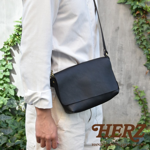 一名男子身穿白色襯衫和米色褲子，肩上斜背著一個黑色皮革郵差包。包包的皮革紋理清晰可見，搭配金屬扣環。背景是灰色的水泥牆和綠色的藤蔓植物。包包右下角印有HERZ品牌標誌和HAND MADE IN JAPAN Since 1973字樣。