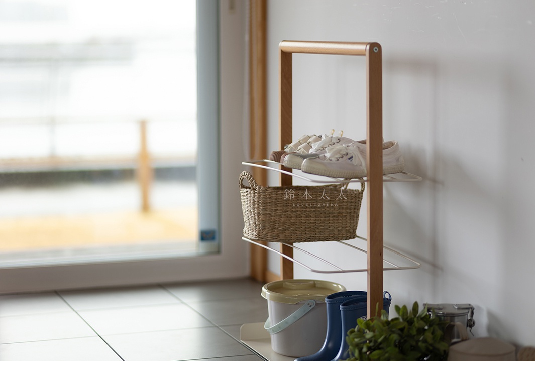 木質框架搭配白色網架的兩層鞋架，上面擺放著一雙白色帆布鞋和一雙深棕色鞋子，中間層有一個編織籃，下方還有一個白色水桶、兩雙藍色雨鞋和一些綠色植物。