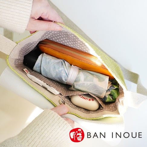 BAN INOUE 品牌標誌旁，黃褐色拉鍊長夾、摺疊傘和白色小肥皂放在淺綠色裂織手提袋中。