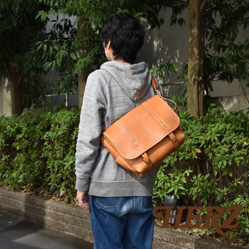 職人手作皮革復古郵差包，採用棕色皮革製成，設計簡約，帶有金屬釘飾和縫線細節，可斜背於肩上，展現復古風格。