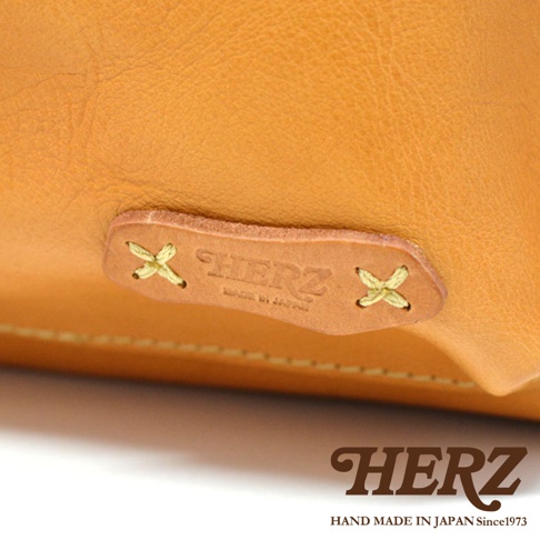 職人手作皮革後背包，標誌為壓印「HERZ」字樣的淺棕色皮革貼片，以黃色線縫合固定在淺棕色皮革表面。