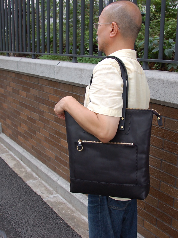 黑色皮革製直式拉鍊托特包，正面有拉鍊口袋和金色拉鍊，側面有金色環扣。