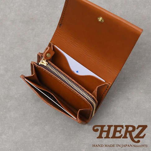 日本品牌HERZ職人手作皮革迷你短夾，採用質感溫潤的棕色天然皮革製成。錢包採雙折開啟設計，內側設有金屬拉鍊零錢袋及多層收納空間，簡潔的構造展現細膩的手工細節，右下角印有HERZ品牌標誌。