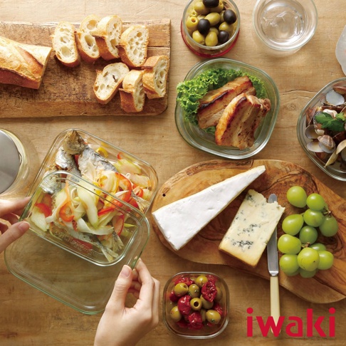 一個透明耐熱玻璃方形保鮮盒，裝有魚和蔬菜，旁邊是麵包、橄欖、烤肉、起司和葡萄。