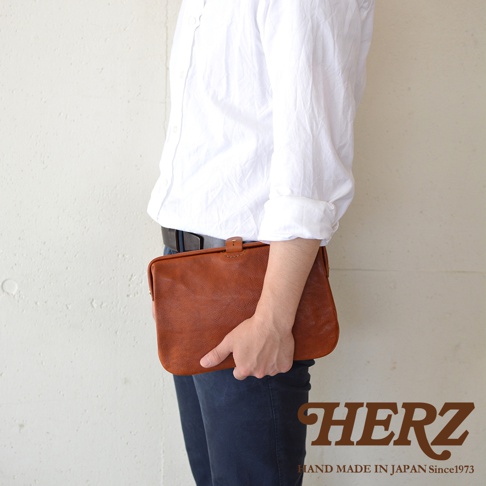 一只手拿著一個棕褐色皮質手拿包，包包表面有自然的紋理，上方有一個皮質搭扣。