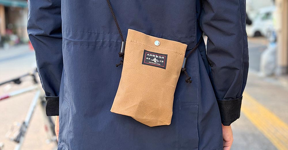 犬印鞄製作所的純綿10號帆布手機包，以褐色帆布製成，配有黑色繩帶，掛在身著深藍色外套的人身上。