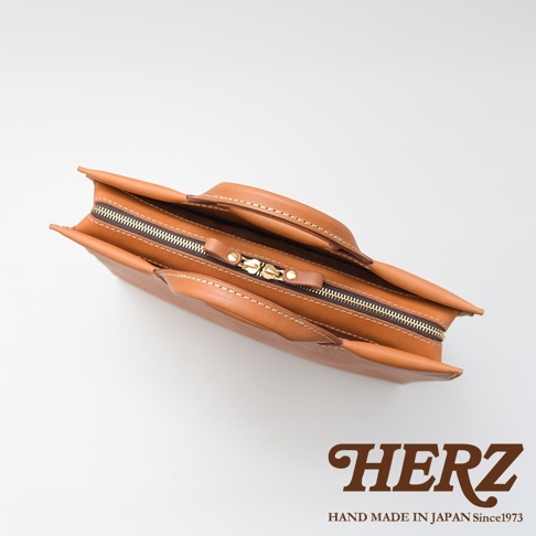 日本HERZ職人手作皮革公事包，頂部特寫，展現皮革的自然紋理與精緻縫線，搭配黃銅色拉鍊與提把，下方印有HERZ品牌Logo與「HAND MADE IN JAPAN Since1973」字樣。