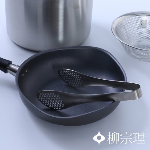 柳宗理不鏽鋼廚具組，包含一個不鏽鋼單柄鍋、一個細網濾網以及置於黑色鑄鐵煎鍋內的洞洞料理夾，整體呈現日式極簡風格，右下角印有柳宗理品牌名稱與商標。