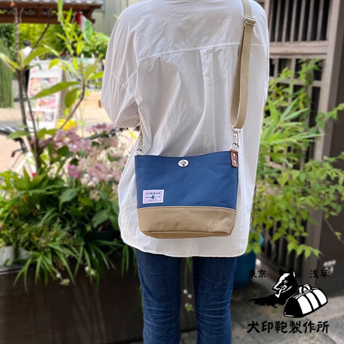 犬印純綿帆布雙色迷你側背包，採用海軍藍與卡其色拼接設計，配有可調式帆布肩帶和金屬扣環。包包正面印有品牌標籤。