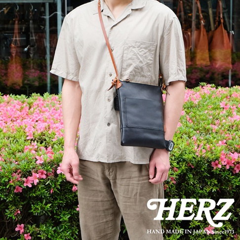 一名男子身穿淺色襯衫和卡其色長褲，斜挎著一個黑色皮革直式隨身單肩包，包包上有一個正面口袋和一個皮質標籤，搭配棕色皮革肩帶。