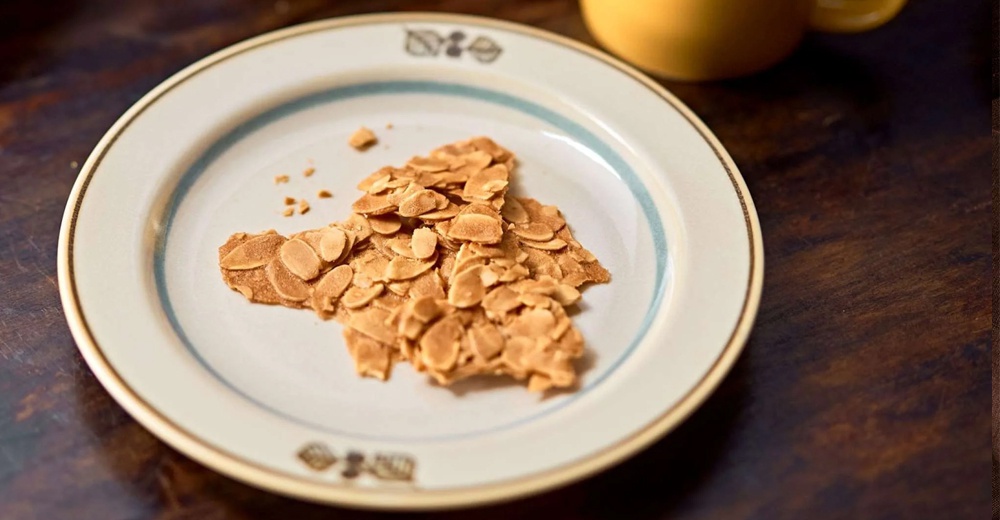 Anyware 美濃燒葉紋復古溫潤甜點盤 M，盤身為米白色，邊緣有藍色與金色雙層細線裝飾，盤底印有淺棕色葉紋圖案。盤中盛放著一片片金黃色的杏仁瓦片點心。