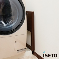 淺米色洗衣機旁，一個深棕色折疊式高腳置物籃靠牆立著，籃框為深棕色，籃板為淺白色。