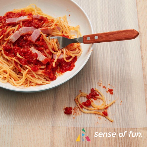 一個白色盤子裝著義大利麵，上面淋有番茄醬和培根，旁邊的叉子有木製手柄。盤子下方是一張淺色、有紋理的保潔墊，上面散落著幾根義大利麵和醬汁。