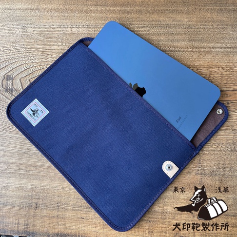 犬印純綿帆布平板收納包，深藍色，可容納一個藍色 iPad。包包外觀為帆布材質，內襯為咖啡色，並有銀色按扣。左側有一個白色方形標籤，印有品牌標誌和文字。