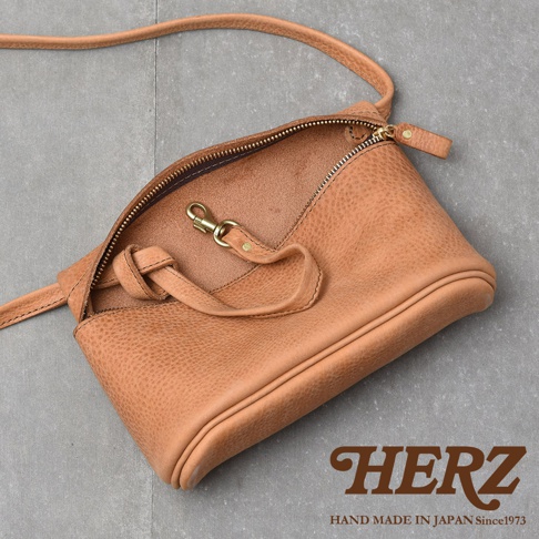 HERZ 手工皮革斜背包，淺棕色，拉鍊半開，露出內部金色掛鉤和綁帶細節，包包表面帶有自然的皮革紋理。