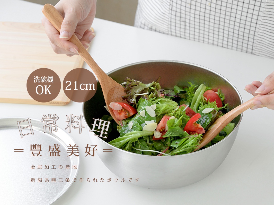 一位女士正用木質湯匙在不銹鋼碗中攪拌沙拉，碗中有生菜、番茄和杏仁片。碗旁有一個木質砧板。碗上印有文字，其中一個圓形標籤顯示「洗碗機 OK」和「21cm」。