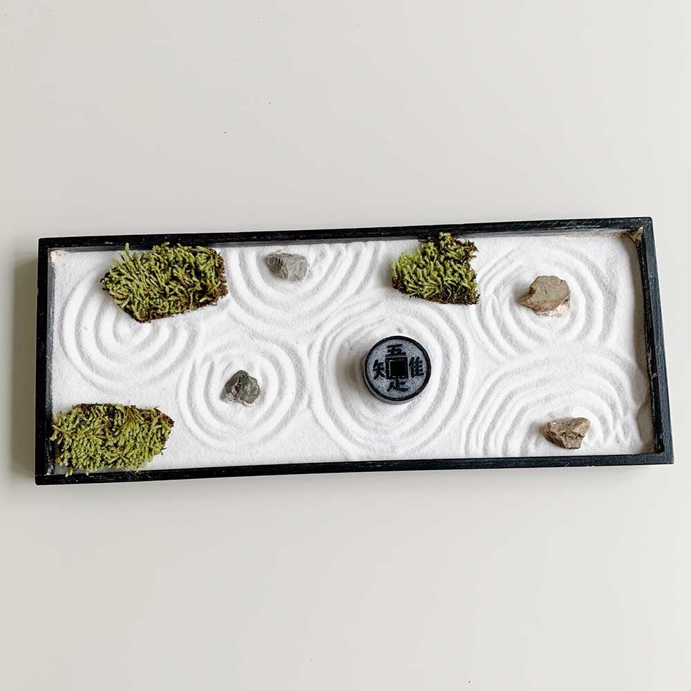 黑色邊框的枯山水模型，內部鋪設白色細砂，砂上耙出同心圓紋路，點綴著數塊灰色石頭和幾叢翠綠苔蘚，中央放置一枚有漢字「知足」的黑色圓形裝飾物。