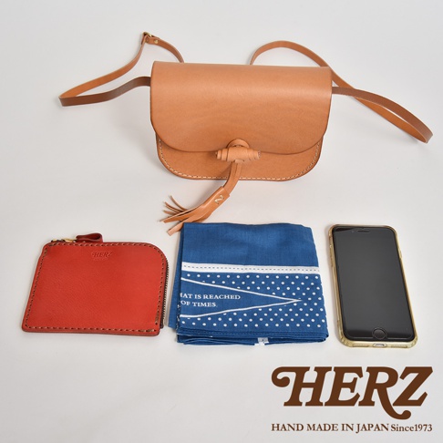 職人手作皮革流蘇側背包，採用原色皮革製成，搭配紅色彩色零錢包、藍色印花手帕及一支手機，展現簡約實用的生活風格。