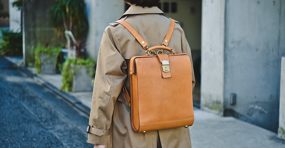 淺棕色皮革製的杜勒斯商務後背包，方形設計，配有金屬鎖扣、提把與皮革肩帶，縫線清晰可見，展現職人手作質感。