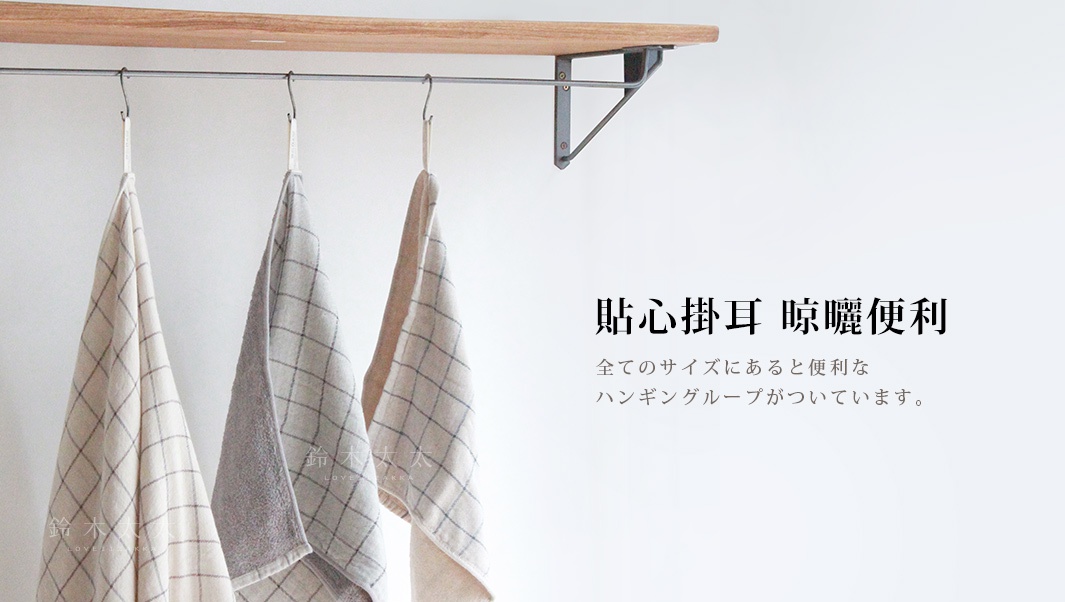 三條GRAPH草木染格紋有機棉毛巾，呈米白色、淺灰色和淺卡其色，帶有細緻的深色格紋圖案，材質柔軟，懸掛於金屬掛架上。