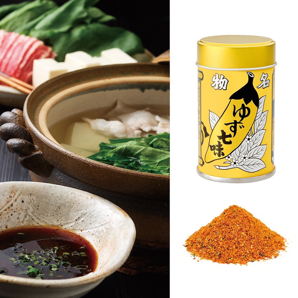 黃色圓罐裝柚子七味粉，罐身印有黑色辣椒和日文「ゆず七味」，下方為一堆橘紅色的七味粉。罐旁為一鍋日式火鍋，內有湯、肉片、豆腐和青菜，另有一碗醬料。