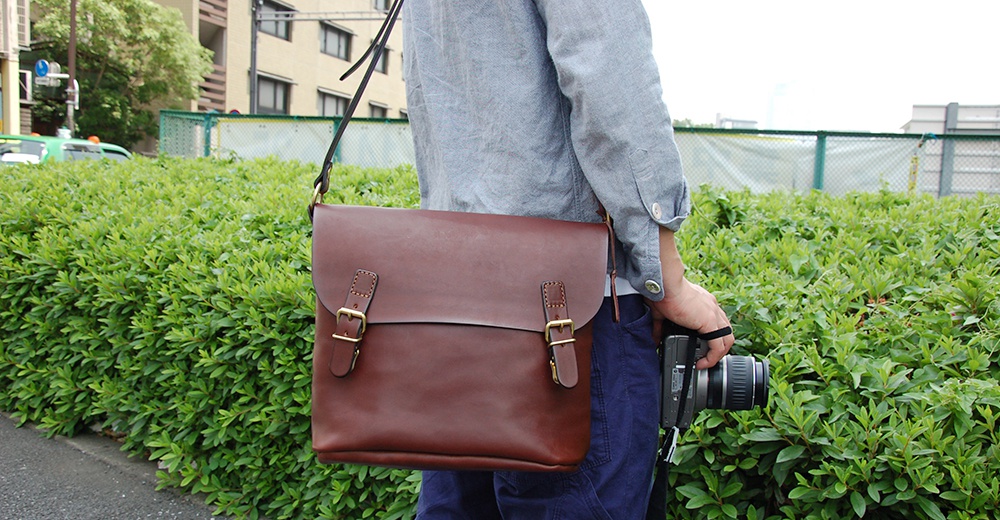日本HERZ職人手作皮革雙扣旅行郵差包，採用Latigo牛皮革製成，搭配雙插釦設計，由一位身穿淺灰色襯衫、藍色長褲的人士攜帶，並手持相機。