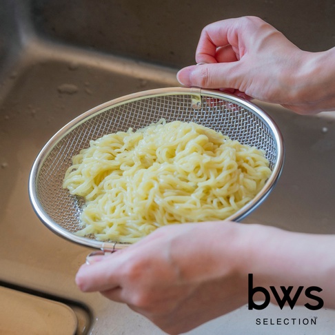 一名女子正在將網篩中的麵條倒入水槽，網篩上印有bws SELECTION的品牌標誌。