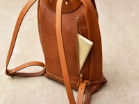 職人手作皮革後背包，色澤為溫暖的棕色，材質為真皮。包體帶有簡約的褶皺設計，側邊有一個拉鍊口袋，內有放置一本淺黃色筆記本。包款配有可調整的皮革肩帶和金屬扣環。