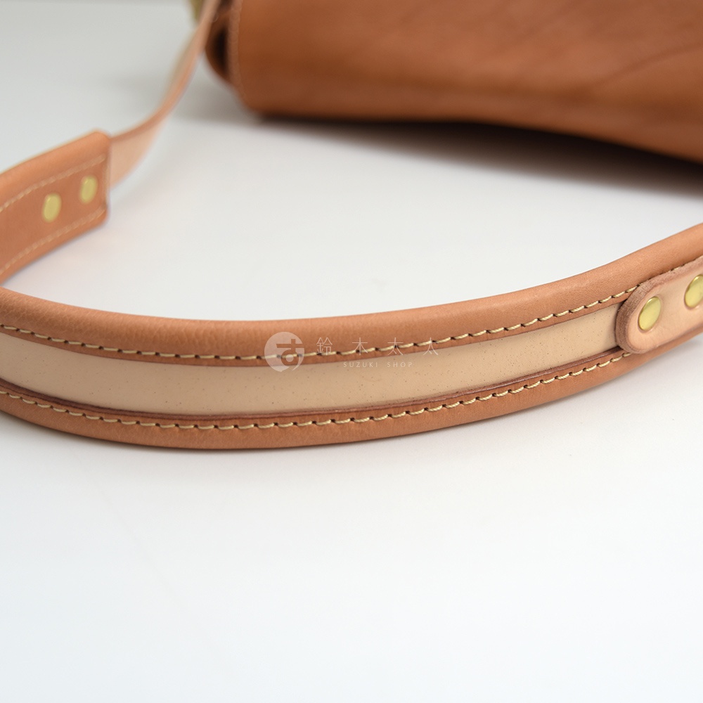 職人手作皮革郵差包的提把特寫，呈現棕褐色皮革和淺棕色皮革拼接，搭配縫線和金屬釘扣，展現優質工藝。