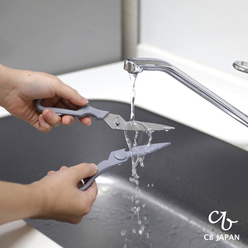 CB JAPAN 廚房剪刀在水龍頭下沖洗，銀色刀刃與灰色手柄，上方一把，下方緊鄰一把，水流向下濺起。