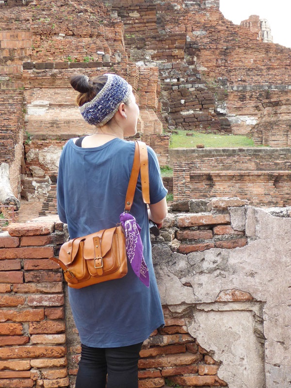 棕褐色皮革製雙口袋肩背包，配有可調式肩帶和兩個帶有搭扣的翻蓋口袋。紫色印花頭巾綁在背包肩帶上。