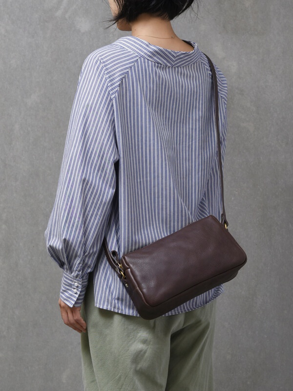 深棕色粒面皮革製成的輕巧拉鍊單肩包，帶有可調節的深棕色皮革斜挎帶和金色金屬配件。