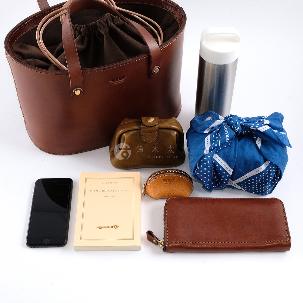 職人手作皮革束口手提包，深棕色皮革，搭配同色系束繩與提把。旁邊擺放一個棕色皮革零錢包、一個棕色皮革長夾、一本淺黃色封面書籍、一個銀色保溫瓶，以及一個藍色印花布包裹的物品。