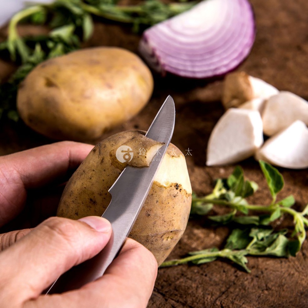 志津匠 Nude+ 削皮刀正在削去一塊馬鈴薯的皮，旁邊有另一塊馬鈴薯、切片的紫洋蔥、菇類塊狀物和香草。