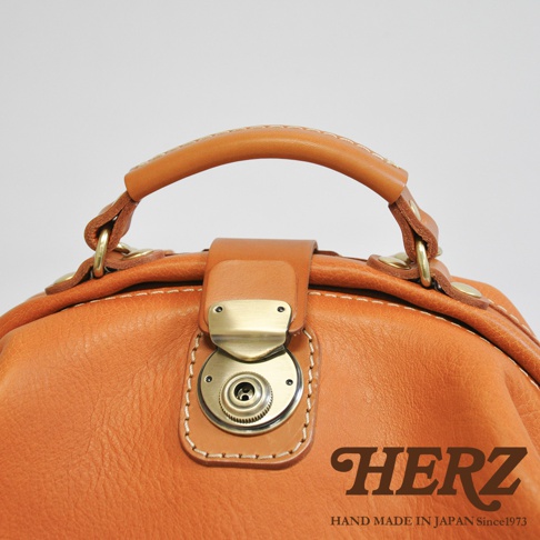 職人手作皮革後背包，採用溫暖的橘棕色皮革，搭配復古金屬旋轉鎖扣與提把，縫線精緻，展現職人細膩工藝。