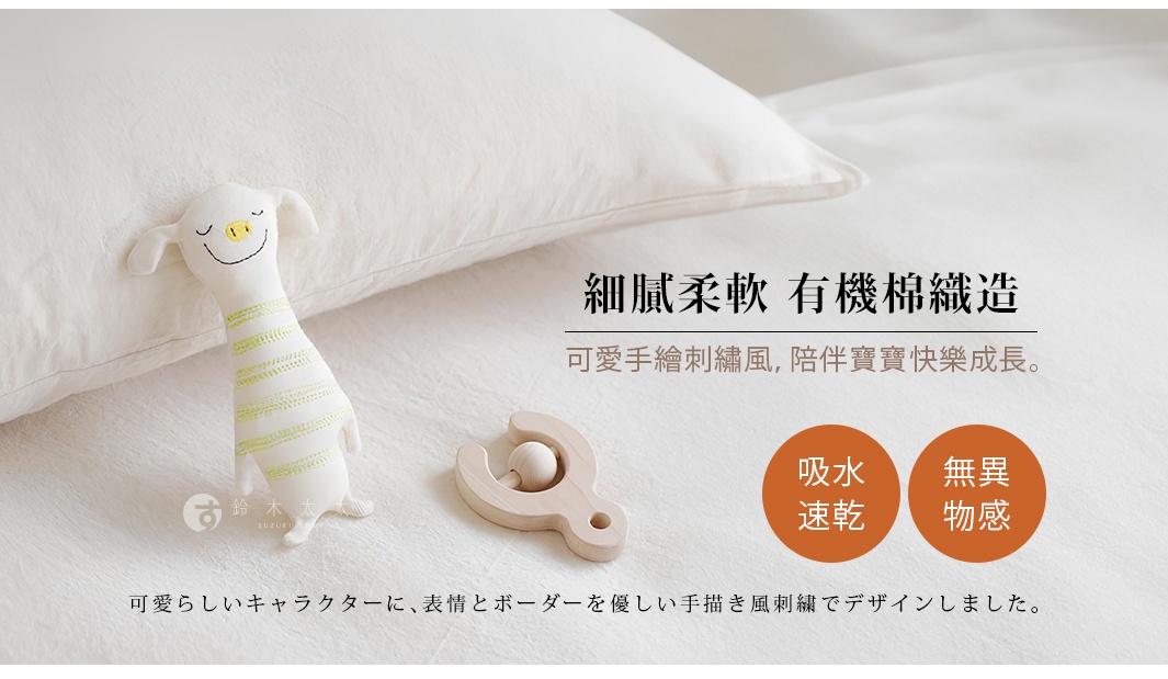 白色有機棉紗布毛巾，搭配綠色條紋的動物造型布偶，及木製搖鈴，置於白色床單上。