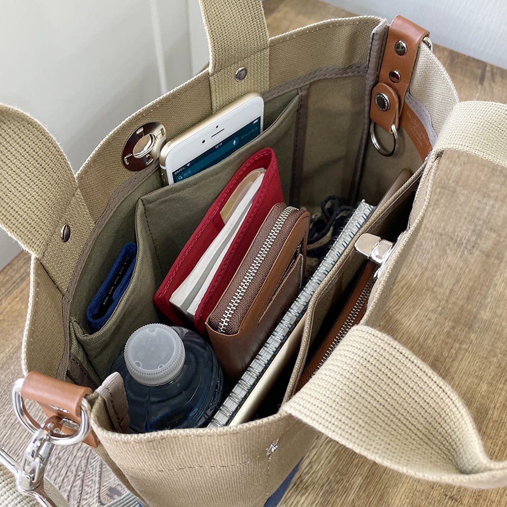 犬印純棉帆布兩用雙色托特包，展示包內物品，包含手機、紅色筆記本、棕色皮夾、筆記本及水瓶，包身為米白色帆布材質，內裡為綠色。