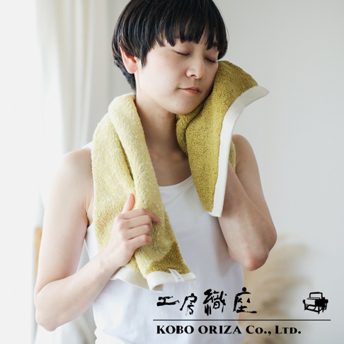 女性用黃綠色棉麻混紡雙色快乾毛巾，一條搭在肩上，另一條擦拭臉部，毛巾邊緣為白色。