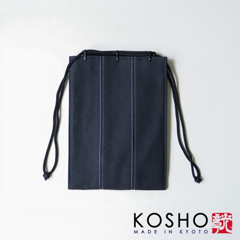 京都KOSHO品牌的一款深藍色帆布信玄袋，採用極簡風格設計，包身中央有兩道垂直的淺色縫線裝飾，頂部配有黑色繩帶與金屬扣件，整體呈現精緻的京都工藝質感，底部右側印有品牌標誌與文字。